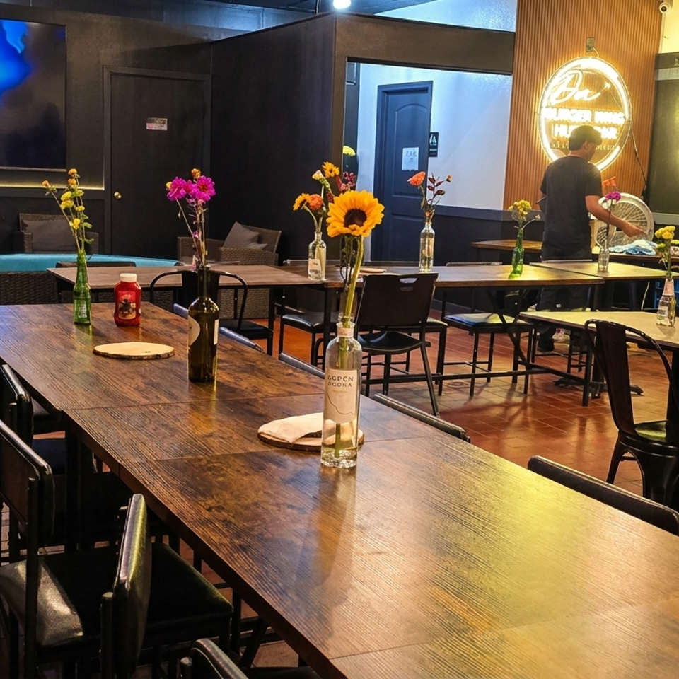 Interior of a restaurant with wooden tables, chairs, and floral centerpieces, with a large TV screen.