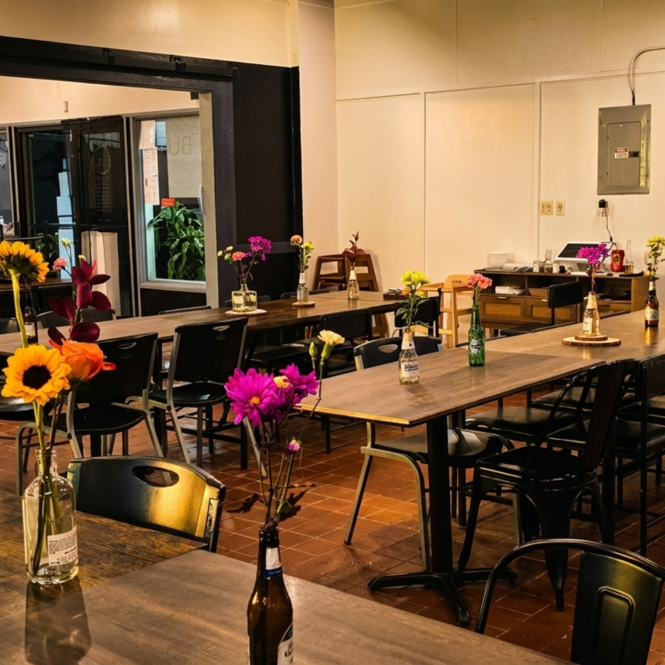 Restaurant interior with long wooden tables, black chairs, and colorful flower arrangements in bottles.