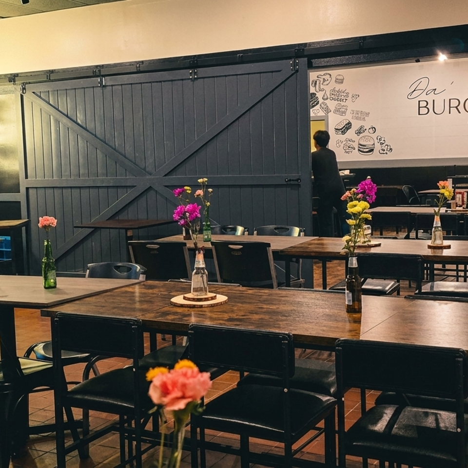 Interior of Da' Burger Wing Hub restaurant with wooden tables, black chairs, and a neon sign.